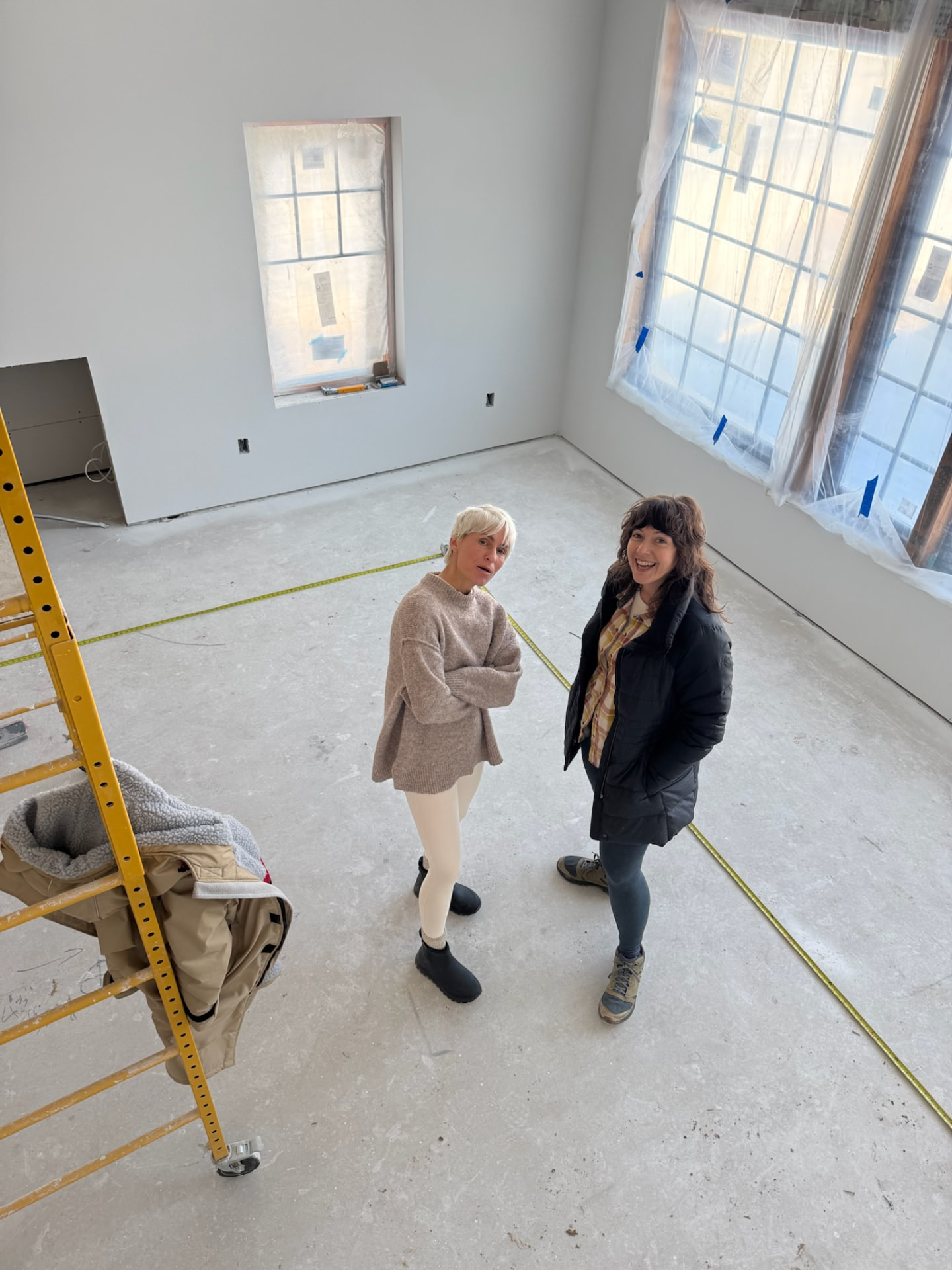 Erika and Stefanie standing in the unfinished great room, tape measure running across the floor
