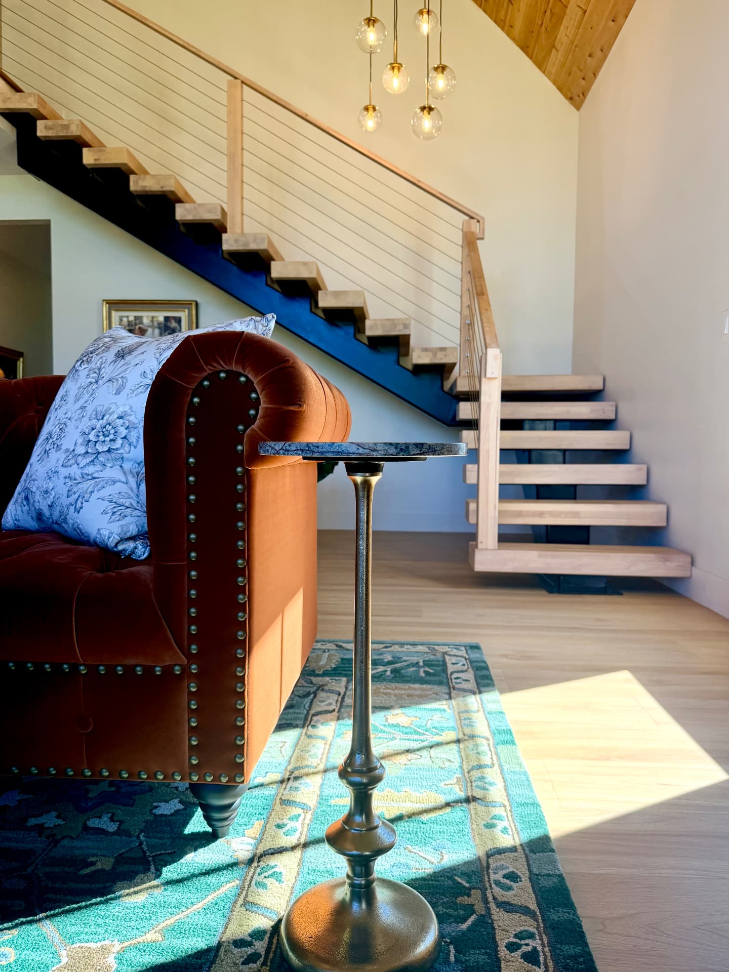 A tufted sofa and bronze drink table in sunlight beside the floating staircase