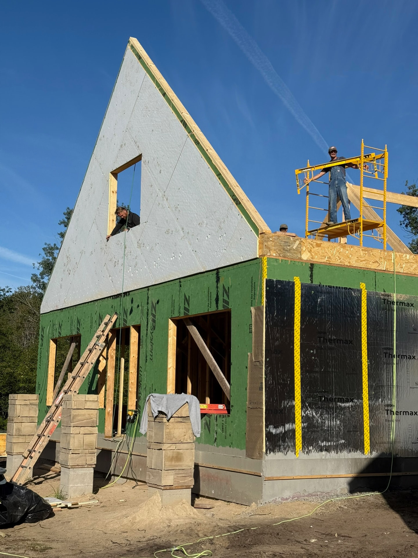 The tall A-frame wall going up, one builder on scaffolding and another framed in the window opening