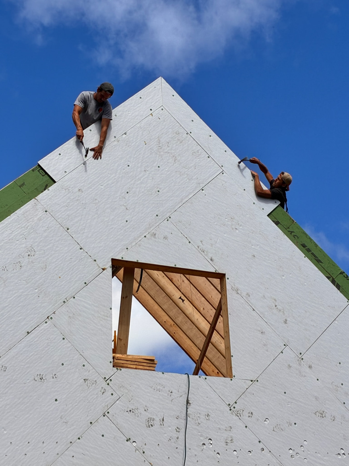 Two builders hammering sheathing at the peak of the roof against a bright blue sky