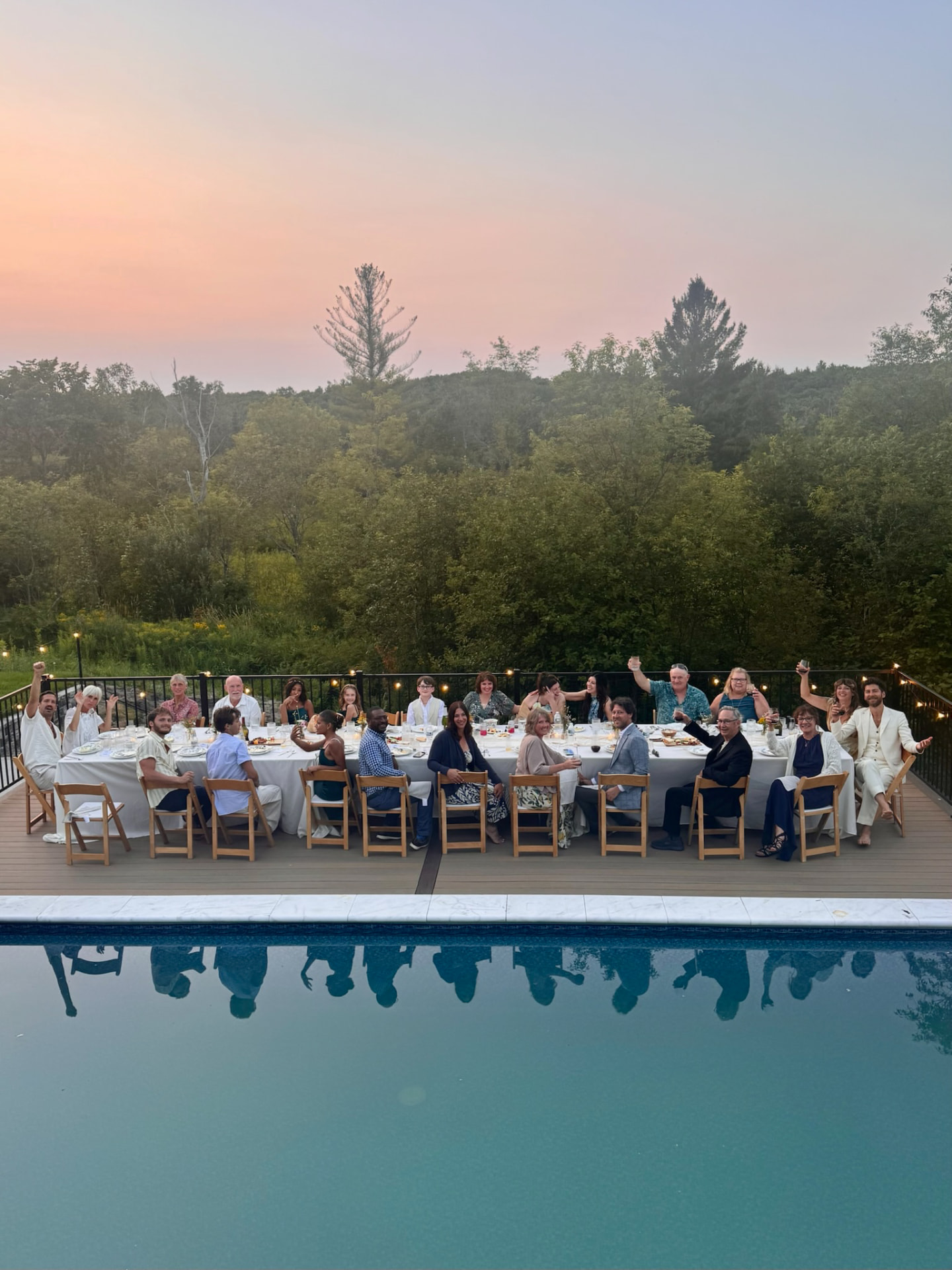 A long table of guests toasting at dusk on the deck, the pool reflecting the gathering below a soft sunset sky